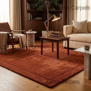 May include: A terracotta-coloured rectangular rug with a darker border, in a living room. The rug is central, with wooden furniture, a leather armchair, and a beige sofa. A wooden side table holds books and a lamp, creating a warm atmosphere.