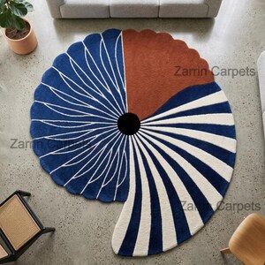 May include: A circular rug with a scalloped edge featuring a central black circle. The design includes a deep blue section with white radiating lines, a solid rust-colored segment, and a section with alternating white and deep blue stripes.