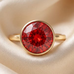 Può includere: Un anello in oro con una pietra preziosa rossa vibrante, rotonda e sfaccettata, montata in una montatura a lunetta liscia. Il gambo dell'anello è in oro lucido e la pietra centrale mostra una brillante tonalità cremisi con sfaccettature.