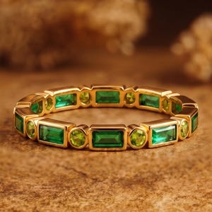 May include: A gold band ring featuring an alternating pattern of rectangular baguette-cut green gemstones and small round-cut green gemstones set in a continuous row around the entire circumference of the metal band.