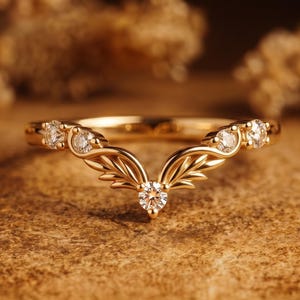 Könnte beinhalten: Ein goldfarbener Ring mit V-förmigem Design. Der Ring verfügt über kleine, klare Edelsteine und einen zentralen, größeren Edelstein. Das Design umfasst stilisierte blatt- oder flügelförmige Elemente, die ein elegantes und zartes Aussehen erzeugen.