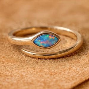 Könnte beinhalten: Ein goldfarbener offener Ring mit einem marquise-geschliffenen Opal-Edelstein. Der Opal zeigt irisierende Blitze in Blau, Orange und Grün. Die Ringschiene ist glatt und poliert, wobei der Edelstein an einem Ende des offenen Designs sitzt.