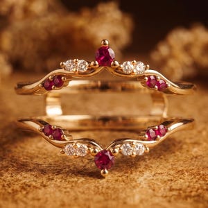 Puede incluir: Un protector de anillo de oro que presenta dos bandas curvas conectadas por un puente. El diseño está acentuado con pequeños rubíes redondos de color rojo intenso y diamantes blancos brillantes engastados a lo largo del marco de metal dorado.