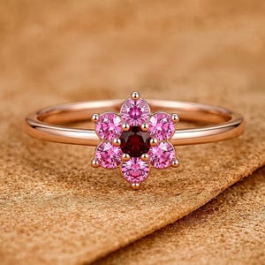 May include: A rose gold ring featuring a flower-shaped cluster setting. The centre stone is a dark red round gemstone, surrounded by six smaller, vibrant pink round gemstones that form the petals of the floral design on a thin metal band.