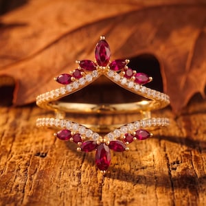 May include: A gold ring guard featuring two chevron-shaped bands adorned with marquise-cut deep red rubies and small round white diamonds. The design includes a central marquise ruby at the peak of each band, surrounded by smaller rubies and diamonds.