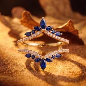 Peut inclure: Une bague double en or composée de deux anneaux en forme de chevron, ornés de saphirs bleus taille marquise et de petites pierres blanches rondes. Le design présente un saphir marquise central au sommet de chaque anneau, entouré de pierres.