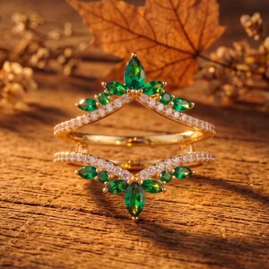 Puede incluir: Un anillo de oro con diseño de doble chevrón, adornado con piedras verdes de corte marquesa y pequeñas piedras redondas transparentes. El anillo tiene una estructura puntiaguda en forma de corona con acentos verdes y transparentes.