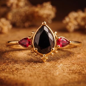 May include: A gold ring featuring a central pear-cut black gemstone flanked by two smaller pear-cut red gemstones. The setting has intricate metal scrollwork details surrounding the stones, creating a decorative frame on a thin gold band.
