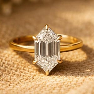 May include: A gold ring with a large, clear, hexagonal-cut gemstone. The ring is set on a neutral-toned fabric, with small white flowers in the background. The ring's band is thin and polished, and the gemstone is faceted to catch the light.