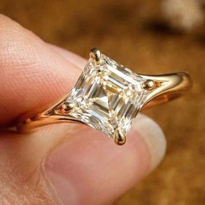 May include: A gold ring featuring a large, square-cut diamond. The ring's band and setting are also gold, with small decorative details on the sides of the diamond. The ring is held between fingers, showcasing its design and sparkle.