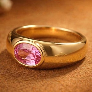 May include: A gold ring with a large, oval-shaped pink gemstone. The ring has a thick band and a polished finish. The gemstone is set in a bezel setting. The background is a warm brown color.