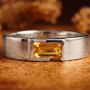 May include: A silver-toned band ring featuring a single rectangular, baguette-cut yellow citrine gemstone set horizontally in the center. The metal band has a smooth, polished finish with a tension-style setting that holds the gemstone securely.