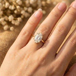 May include: A gold ring with a large, emerald-cut diamond. The ring is set on a delicate gold band. The diamond is clear and reflects light. The ring is on a person's finger.