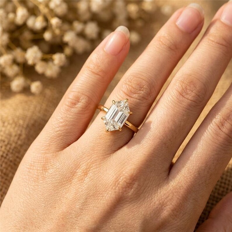 May include: A gold ring with a large, emerald-cut diamond. The ring is set on a delicate gold band. The diamond is clear and reflects light. The ring is on a person's finger.