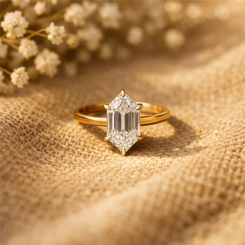 May include: A gold ring with a large, clear, hexagonal-cut gemstone. The ring is set on a neutral-toned fabric, with small white flowers in the background. The ring's band is thin and polished, and the gemstone is faceted to catch the light.