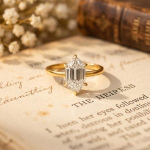 May include: A gold ring with a large, clear, hexagonal-cut gemstone. The ring is set on a vintage book page with the word "HEIRESS" visible. Delicate white flowers are in the background.