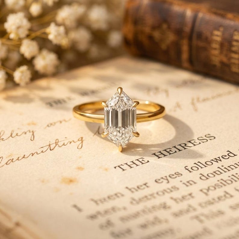 May include: A gold ring with a large, clear, hexagonal-cut gemstone. The ring is set on a vintage book page with the word "HEIRESS" visible. Delicate white flowers are in the background.
