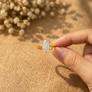 May include: A gold ring with a large, clear, emerald-cut gemstone. The ring is held by a hand against a neutral background, with soft lighting. The ring's band is a simple, polished gold.