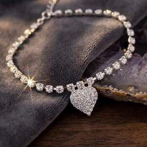 May include: A silver-toned necklace with a heart-shaped pendant. The necklace features a delicate chain adorned with numerous clear, sparkling crystals. The heart pendant is fully encrusted with smaller crystals, creating a dazzling effect. The necklace is displayed on a dark gray surface.