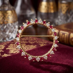 May include: A gold-toned brooch in a circular design, adorned with alternating red and clear gemstones. The brooch is set against a deep red velvet surface, with a gold pin clasp. The gemstones are round and evenly spaced around the circle.