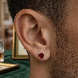 May include: A square-cut, deep red gemstone stud earring set in a silver-toned metal prong setting. The earring is positioned in the earlobe, featuring a faceted surface that reflects light, showcasing a vibrant crimson hue and a classic four-prong design.