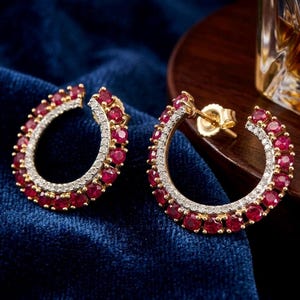 May include: A pair of gold hoop earrings with a double row of small, round, red gemstones and a row of small, clear gemstones. The earrings are set against a dark blue background.