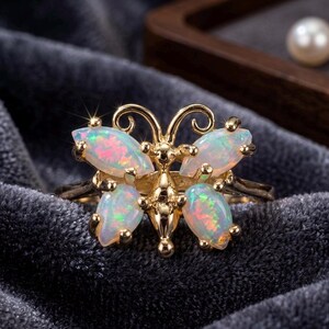 May include: A gold-colored ring featuring a butterfly design. The butterfly's wings are made of oval-shaped opals with iridescent colors of pink, blue, and green. The ring is set against a dark gray velvet background.