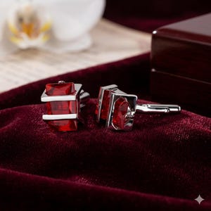 May include: A pair of silver cufflinks with a rectangular red gemstone center. The cufflinks have a modern design with a polished finish, set against a deep red velvet surface.