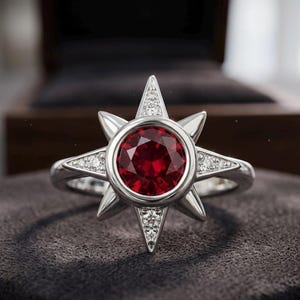May include: A silver ring featuring a round, deep red gemstone set in the center of a starburst design. The starburst points are accented with small, clear, sparkling stones, creating a bright contrast against the polished silver metal band.