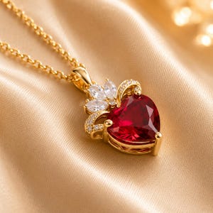 May include: A gold necklace featuring a heart-shaped red gemstone pendant. The pendant is accented with small, clear marquise-cut stones arranged in a crown-like setting above the heart, with additional tiny clear stones along the gold frame.