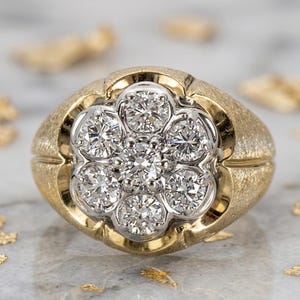 May include: A gold ring with a cluster of diamonds in a flower-like arrangement. The ring has a textured band and a white gold setting for the diamonds. The diamonds are round-cut and sparkle brightly.