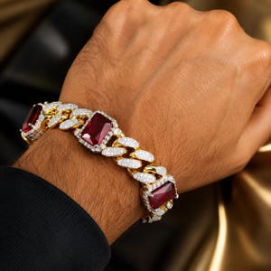 May include: A gold-toned cuban link bracelet featuring three large, rectangular, deep red gemstones. Each gemstone is surrounded by a halo of small, clear, sparkling stones. The links of the chain are also encrusted with small, clear, sparkling stones.
