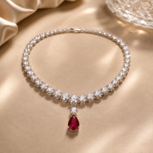 May include: A silver necklace with a series of round, clear stones encircling the neck. A teardrop-shaped, red gemstone pendant hangs from the center. The necklace is displayed on a gold-colored satin surface.