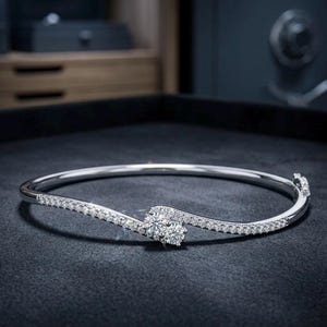 May include: A silver bracelet with a wave design. The bracelet features a large, round, clear gemstone at the center, surrounded by smaller, clear gemstones. The bracelet is set against a dark background.