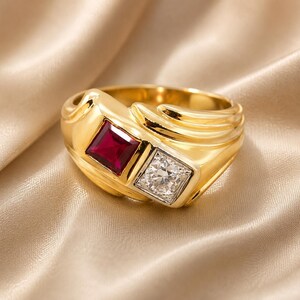 May include: A gold ring featuring a square-cut deep red ruby and a square-cut clear white diamond set side-by-side. The band has a polished gold finish with a series of three raised, curved ridges extending along one side of the ring shank.