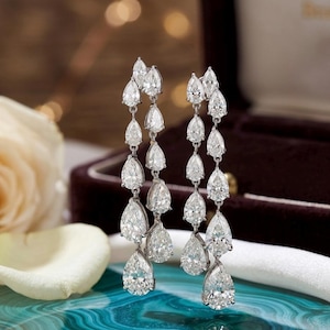 May include: Elegant dangling earrings featuring multiple pear-shaped diamonds. The diamonds are arranged in a cascading design, creating a glamorous and eye-catching look. The earrings are set against a blurred background, highlighting the sparkle of the gemstones.