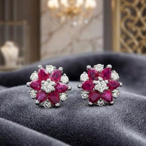 May include: A pair of flower-shaped stud earrings. Each earring features a cluster of deep red gemstones surrounded by smaller, clear gemstones. The earrings are set in a silver metal.