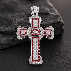 May include: A silver-toned cross pendant featuring a central circular design, adorned with numerous small, clear white stones and vibrant red stones arranged in rectangular patterns across the surface, including a matching bail at the top.