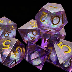 May include: Set of seven polyhedral dice with a shimmering, iridescent effect. The dice are transparent with a purple hue and gold numbering. Ideal for tabletop games, the dice are shown against a black background.