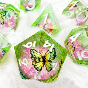Pode incluir: Conjunto de dados poliédricos com um design verde translúcido. Cada dado apresenta uma borboleta e detalhes de glitter rosa. Os números são brancos e os dados estão sobre uma superfície branca e fofa. Para jogos de tabuleiro.