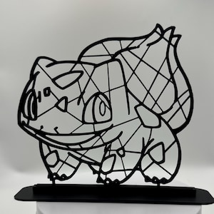 May include: Black metal Bulbasaur figure. The figure is a stylized outline of the Pokémon character, mounted on a rectangular base. The design features geometric shapes within the outline, creating a unique aesthetic. The figure is displayed on a white surface.