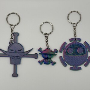 May include: Three keyrings with silver rings. The keyrings feature designs of a skull with a moustache and crossbones, a cartoon character with a hat, and a circular design with a face. The keyrings have a purple and blue iridescent finish.