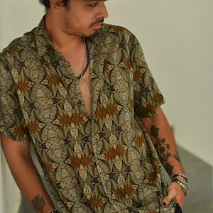 May include: A brown fedora hat and a short-sleeved shirt with a geometric pattern in brown, green, and blue. The person is wearing silver bracelets and a black necklace. The shirt is unbuttoned.