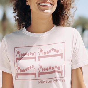 May include: White t-shirt with a pink graphic of caterpillar-like figures on exercise benches. The text "Pilates Mom" is printed below the graphic. The shirt is made of a soft material.