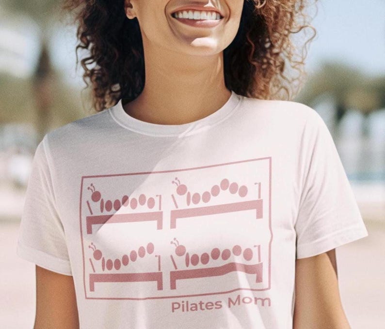 May include: White t-shirt with a pink graphic of caterpillar-like figures on exercise benches. The text "Pilates Mom" is printed below the graphic. The shirt is made of a soft material.