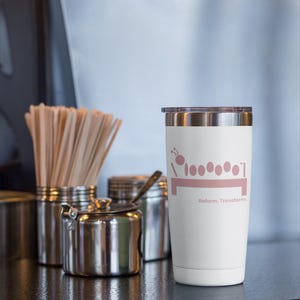 May include: A white insulated tumbler with a silver rim and lid. The tumbler features a pink graphic of a caterpillar and the words "Reform. Transform." on the front. Other stainless steel containers are in the background.