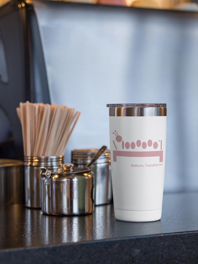 May include: A white insulated tumbler with a silver rim and lid. The tumbler features a pink graphic of a caterpillar and the words "Reform. Transform." on the front. Other stainless steel containers are in the background.
