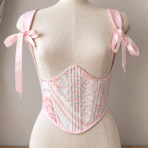 May include: A corset top featuring a light pink and pale blue checkered pattern with white accents. The garment has a pointed hemline, structured vertical seams, and wide satin ribbon shoulder straps that tie into bows at the front shoulders.