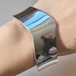 Può includere: Un bracciale a polsino largo, color argento, con un design curvo. Il metallo lucido riflette la luce, creando un aspetto luminoso e brillante. Il bracciale è indossato su un polso, mostrando il suo stile minimalista e moderno.
