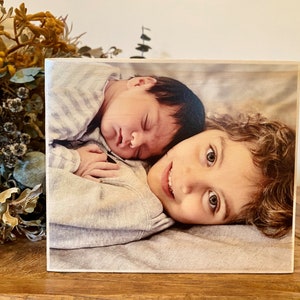 May include: A close-up photo of two siblings. An older child with curly brown hair is lying down with a younger baby sibling sleeping on their chest. The baby is wearing a striped shirt and the older child is wearing a grey shirt.
